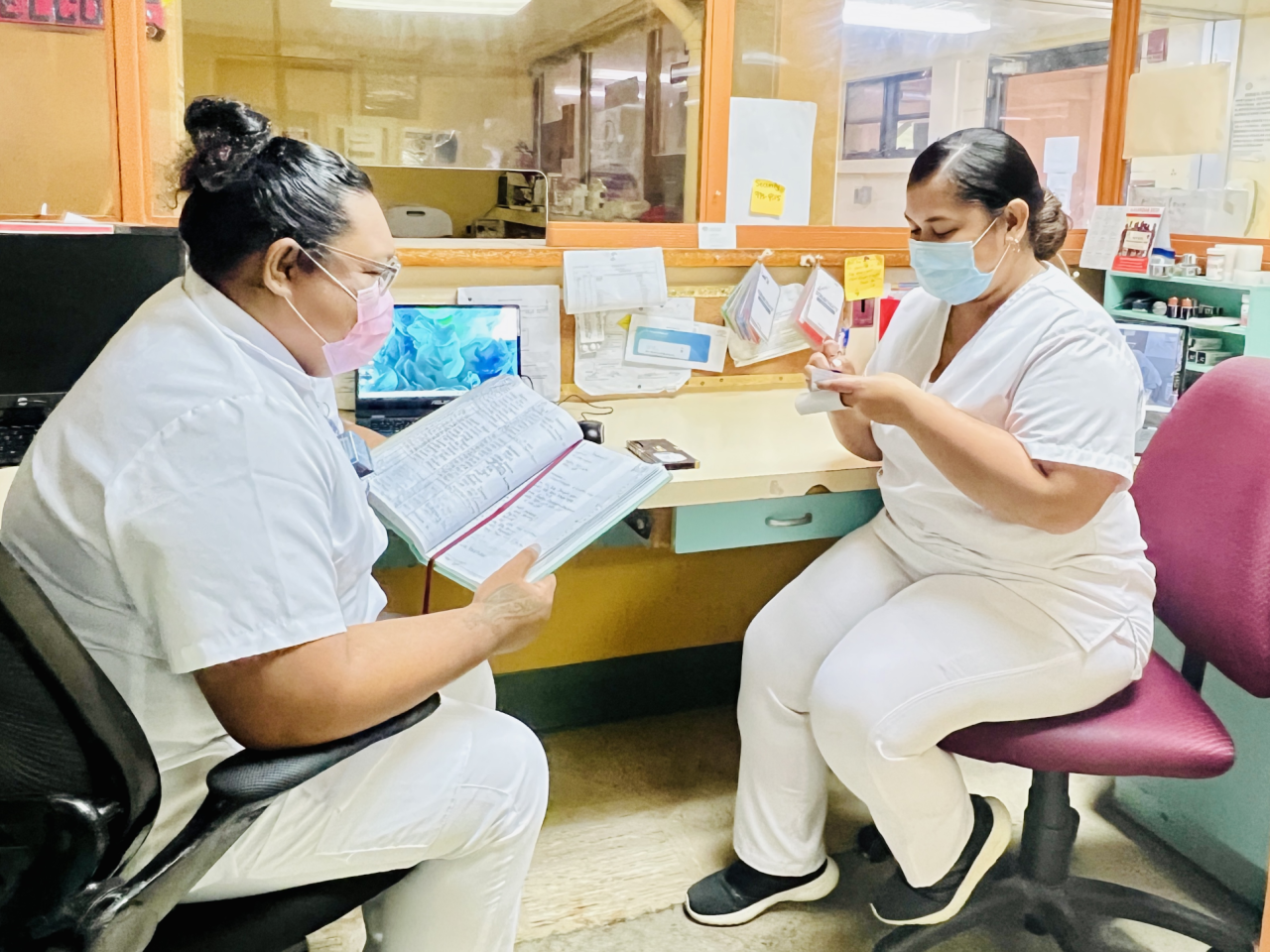 Two women nurses with face masks sitting on chairs inside a hospital reception counter: one woman with a record book in her hands and the other writing down on a piece of paper