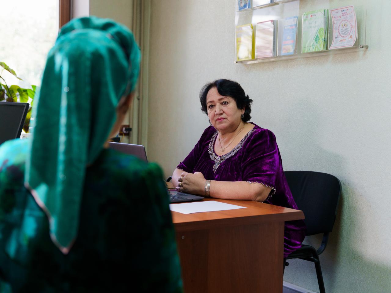 A woman in a purple dress behind a desk speaking to a woman with a green head covering
