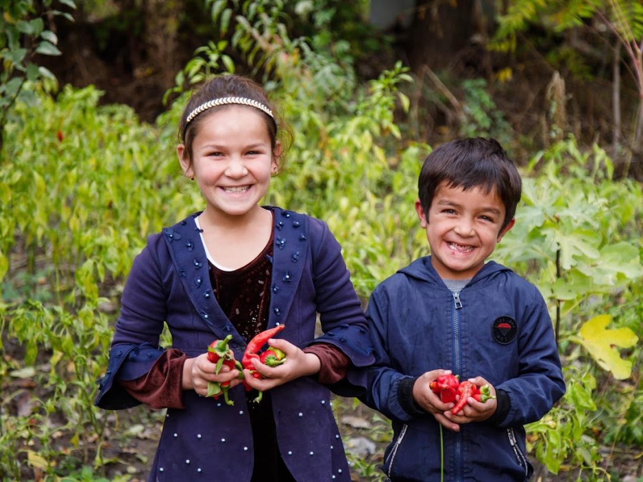 A girl and a boy smile in a garden holding vegetables