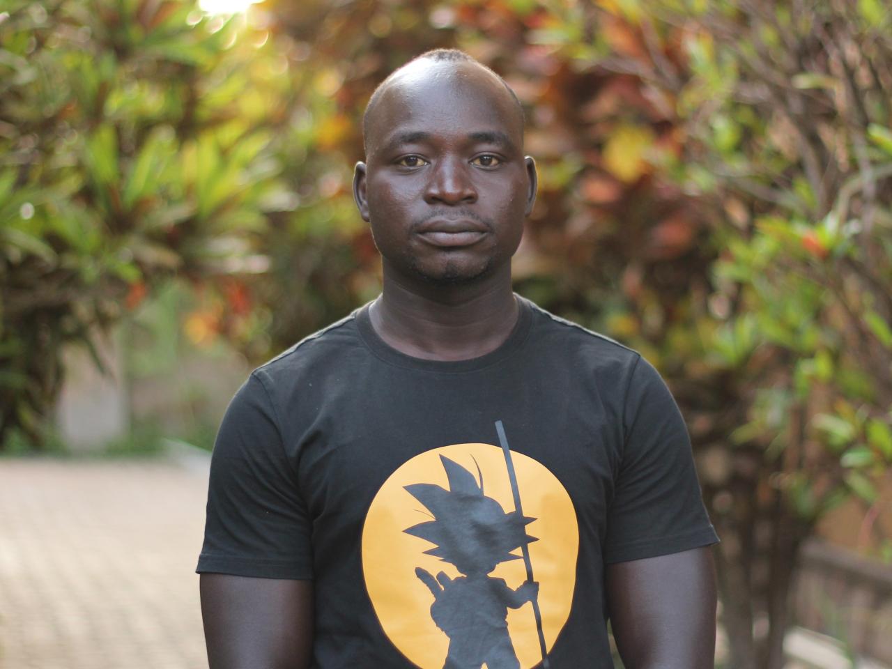 A man wearing a black t-shirt stands outside and looks into camera