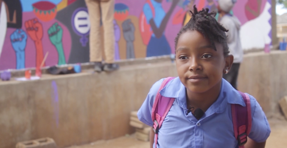 A young girl in school shirt stands in front of mural