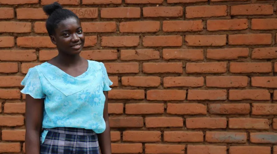 A girl in a blue shirt and checked skirt stands in front of brick wall smiling