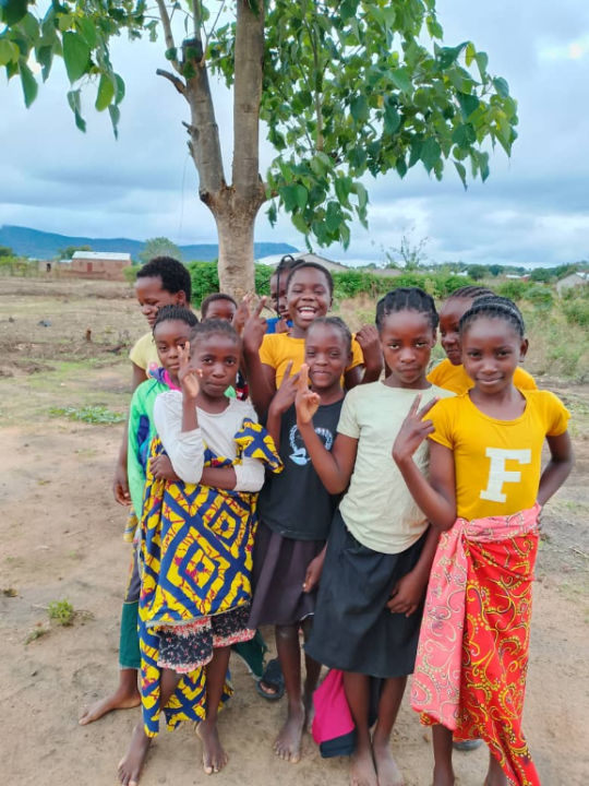 A group of girls aged between 12 and 15 years pose for a photo under a tree with a beautiful scenery of hills behind them. The girls are smiling with some raising fingers in V-shaped peace symbol.