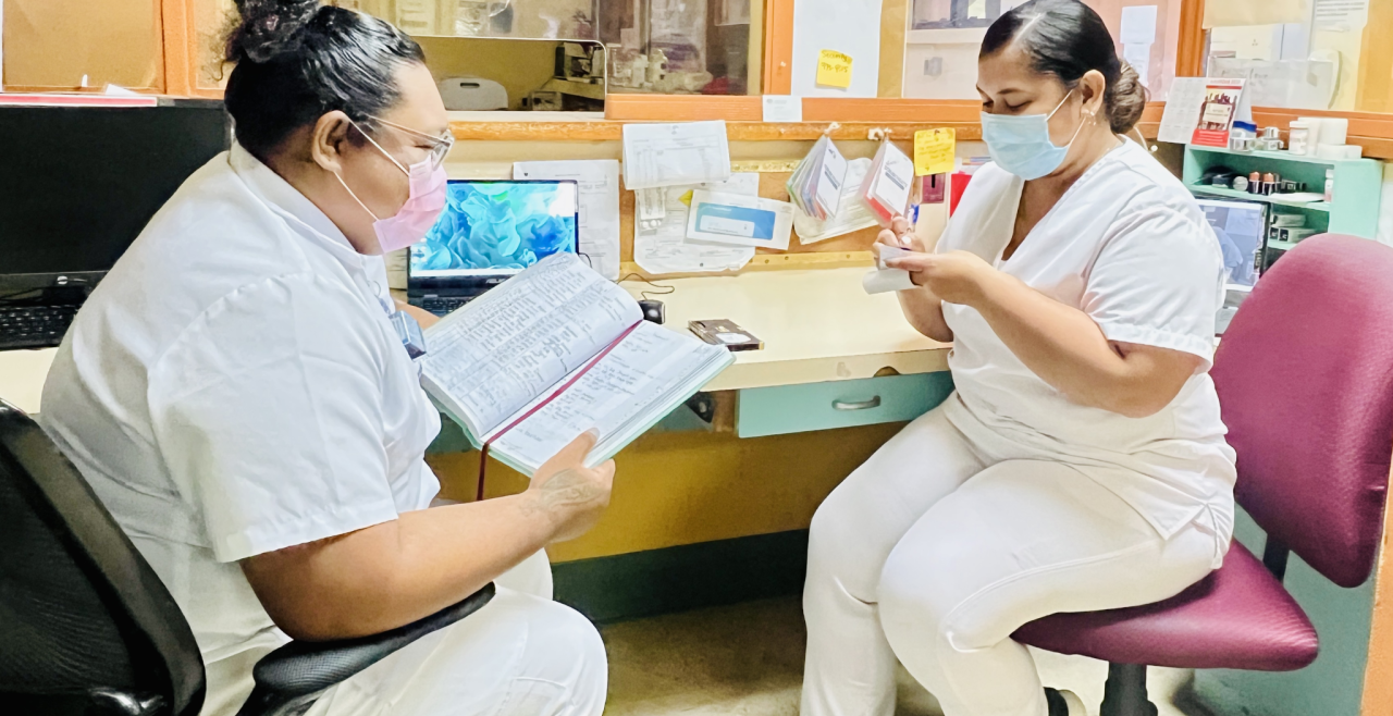 Two women nurses with face masks sitting on chairs inside a hospital reception counter: one woman with a record book in her hands and the other writing down on a piece of paper